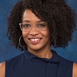 Tiana, an African American woman, sits smiling at the camera. She is wearing a sleeveless navy blue blouse, silver hoop earrings, and glasses with translucent beige frames.