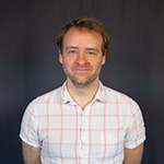 Paul Ellison smiles at the camera. He has short blonde hair and blue eyes and is wearing a white and red button-up shirt.
