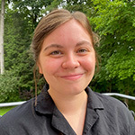 Emmi Litsenberger sits on a porch in front of a grassy lawn and trees. She is smiling and wearing a gray button-up shirt.