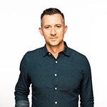 Dean Sell stands in front of a white background. He is wearing a dark button down shirt.