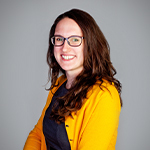 Tasha Rosenbaum smiles at the camera. She has brown hair and is wearing glasses, a yellow cardigan, and a blue sweater.