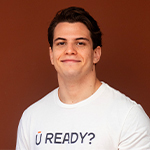 Gabriel Garnier smiles at the camera. He is wearing a white t-shirt that says “U READY?” and standing against a brown backdrop.