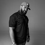 Kevin Hall stands and looks at the camera. He has a beard and is wearing a baseball cap, a collared short-sleeve shirt, and jeans.