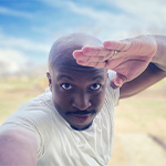 Chancler Haynes stands outside on a sunny day. He is wearing a white t-shirt and saluting at the camera.