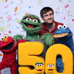 Chris Sassano stands with Oscar the Grouch, Cookie Monster, Elmo, and a puppet shaped like the number 50 with confetti behind them.