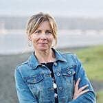 A blonde woman wearing a denim jacket over a navy top and black skirt stands with her arms crossed. She is outdoors in front of a river with mountains far in the background.