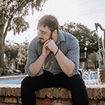 A man with short, dark, curly hair in a blue button down shirt and wireframe glasses looking to the side while sitting in front of a fountain.