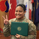 Anjelica Asejo smiles at the camera holding a certificate. She is wearing her Army uniform with a row of flags behind her as she holds her fingers in a peace sign.