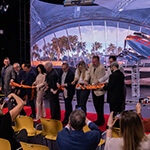 A group of eight men and two women stand on a red carpet in front of a large screen displaying an airplane and palm trees. They are each holding a pair of scissors and cutting a long orange ribbon. The seven audience members in front of them are photographing them with cell phones.