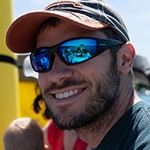 A man wearing a blue t-shirt, a blue and red baseball cap, and sunglasses with polarized lenses sits on a boat on a sunny day. He is smiling at the camera and there is a brunette woman and a blonde child sitting out of focus behind him.