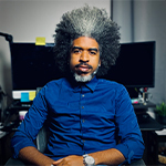 Bill Hudson sits in an office chair in front of a desk with two computer monitors. He has black and gray hair and wears a blue button-up shirt.