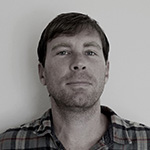 A man in a plaid shirt stands against a plain wall, looking directly at the camera with a very slight smile.