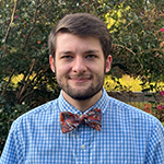 Noah White smiles at the camera in front of a row of green bushes with pink flowers. He is wearing a blue shirt and red bowtie.