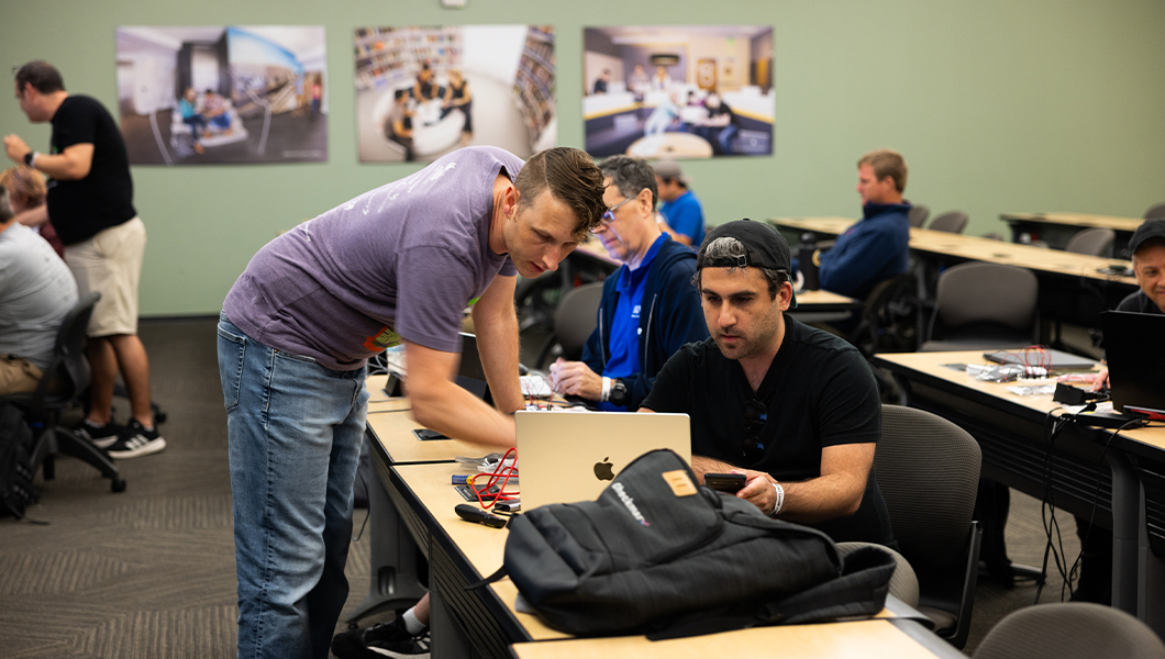 Two students face a laptop on a table, one siting in a chair and one standing over the table. Several other students sit at desks around the room.