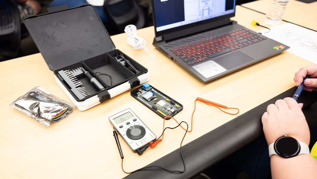 A student sits at a table with a laptop and circuit board soldering kit on a table in front of them. They are wearing a white watch and holding a blue pen.