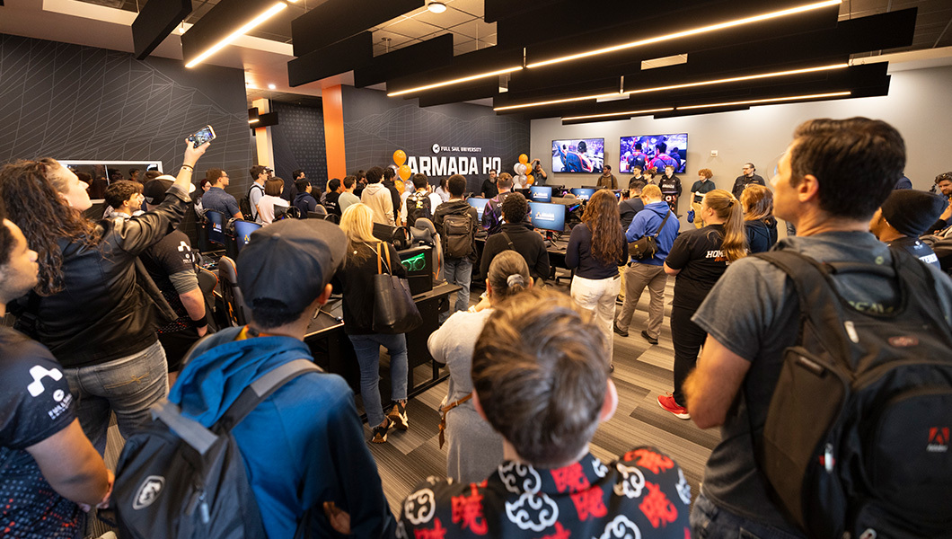 A crowd gathers inside Armada HQ with gaming consoles, TV screens, and an Armada HQ sign framed by orange and silver balloons.