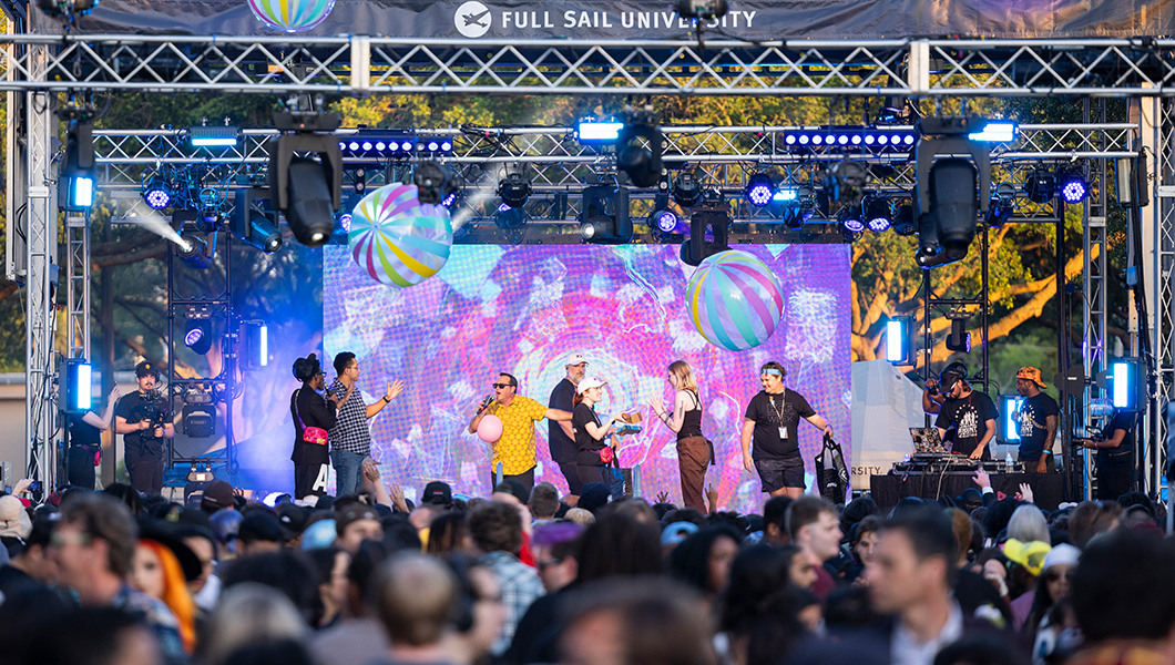 A large crowd is gathered in front of an outdoor stage. Many people are standing on the stage against a multi-colored screen, with one person wearing a yellow shirt that is holding a microphone and addressing the crowd.