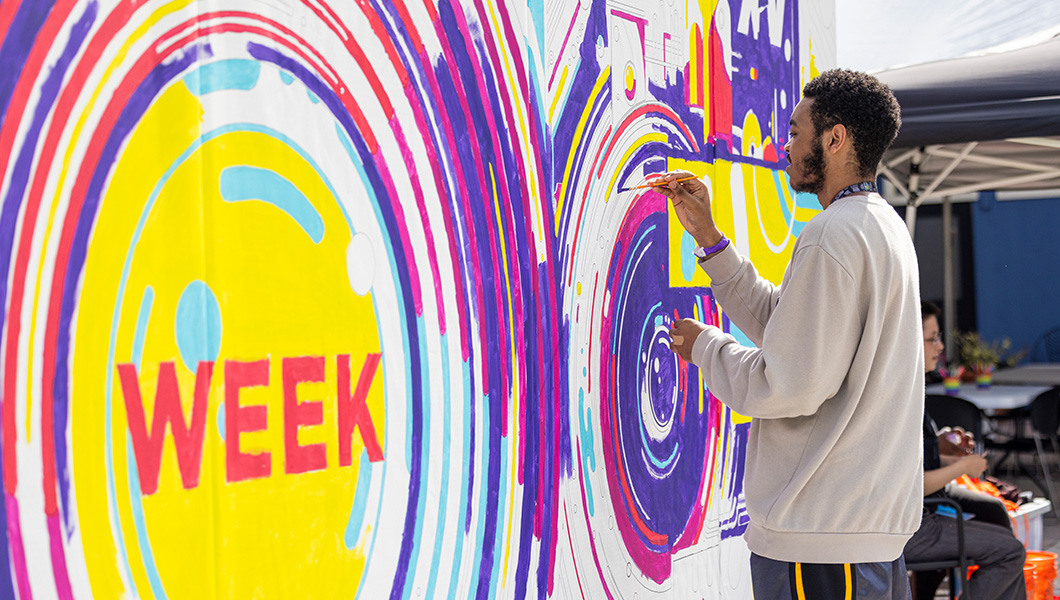A student wearing a gray sweater paints a purple line on a large mural.