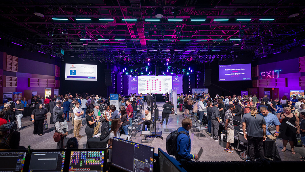 A wide shot of a crowded arena with many booths set up on the floor. Many large screens depict different images in the background. A soundboard sits in the foreground.