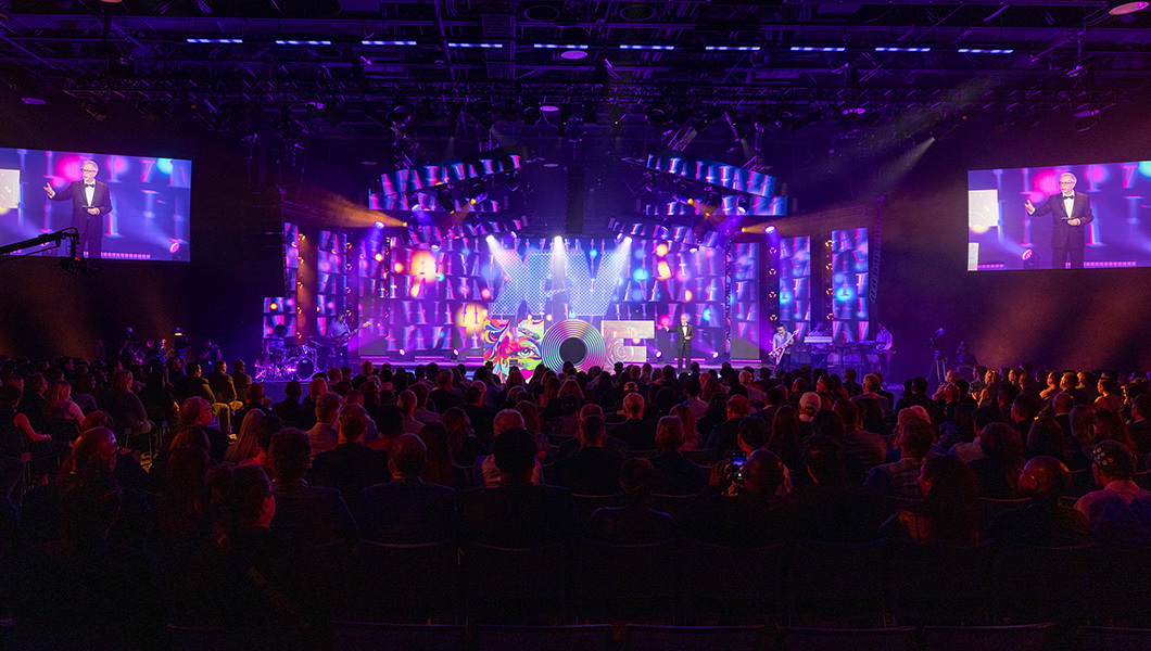 A large crowd is seated in front of a raised stage. A large, multi-colored sculpture of the letters “HOF” sit atop the stage in front of a purple screen. A man wearing a black suit stands on the stage and is pictured in the screens beside the stage.