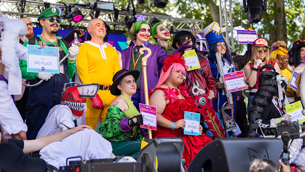 Several cosplayers pose for a picture on an outdoor stage. They are holding signs denoting their placement in the competition.