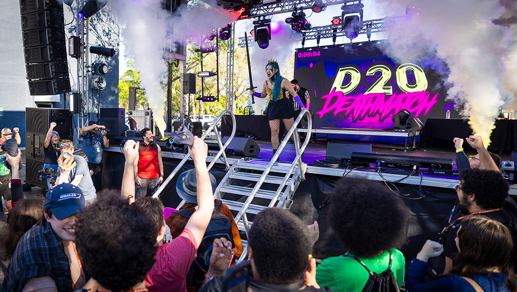 A crowd of students cheer as the D20 Deathmatch takes place on a stage. The host wears a costume and carries a sword.