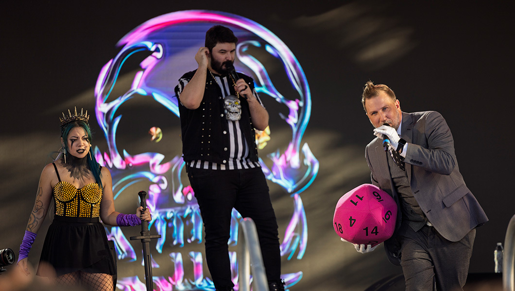 Three hosts in costumes are onstage and one holds a giant 20-sided die. A skull graphic is displayed onscreen behind them.