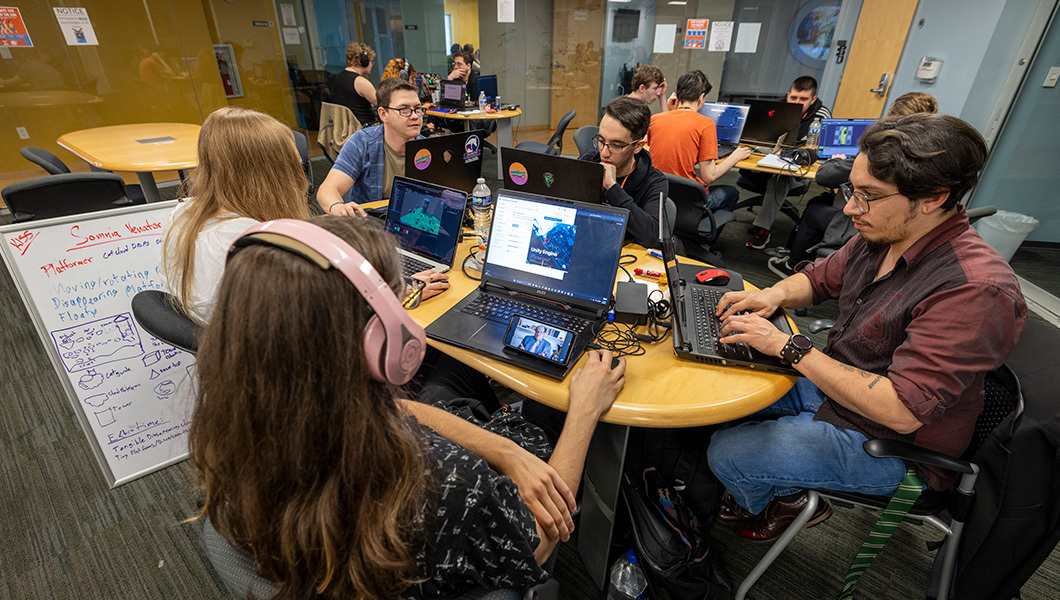 In a classroom, teams of students sit at tables on their laptops.