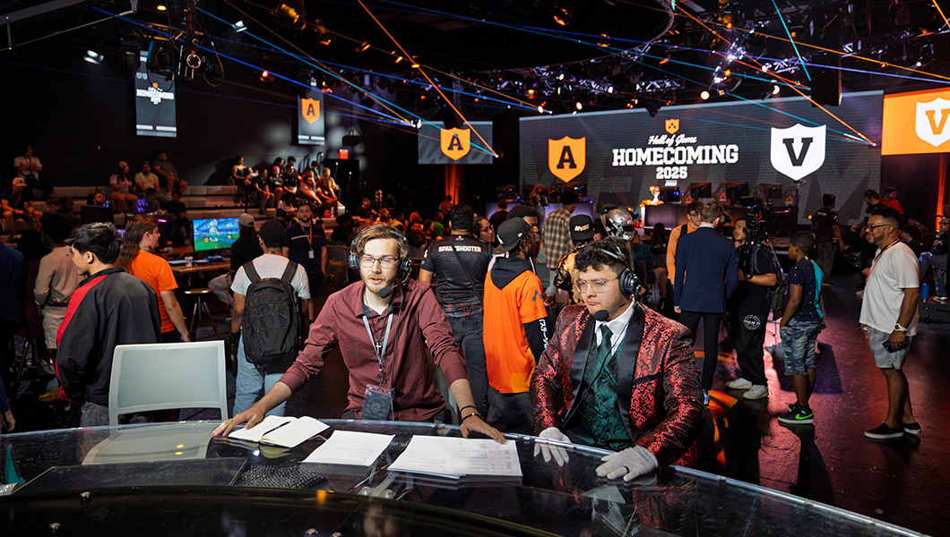 Two hosts wearing headsets share commentary in the Fortress. A crowd of audience members is behind them.