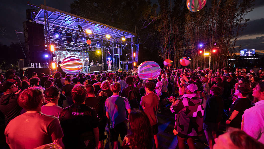 A crowd of students gathers around a stage with live music and lights. Inflatable beach balls are being passed among the crowd.
