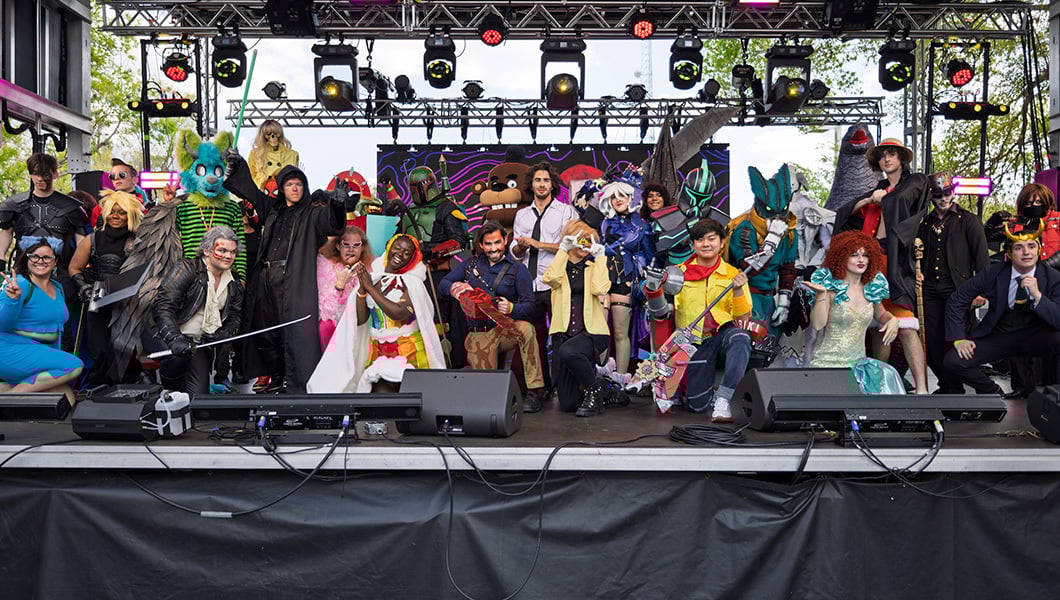 A group of students in cosplay pose on a stage, featuring an array of characters such as Bill Cipher, Boba Fett, and Loki.