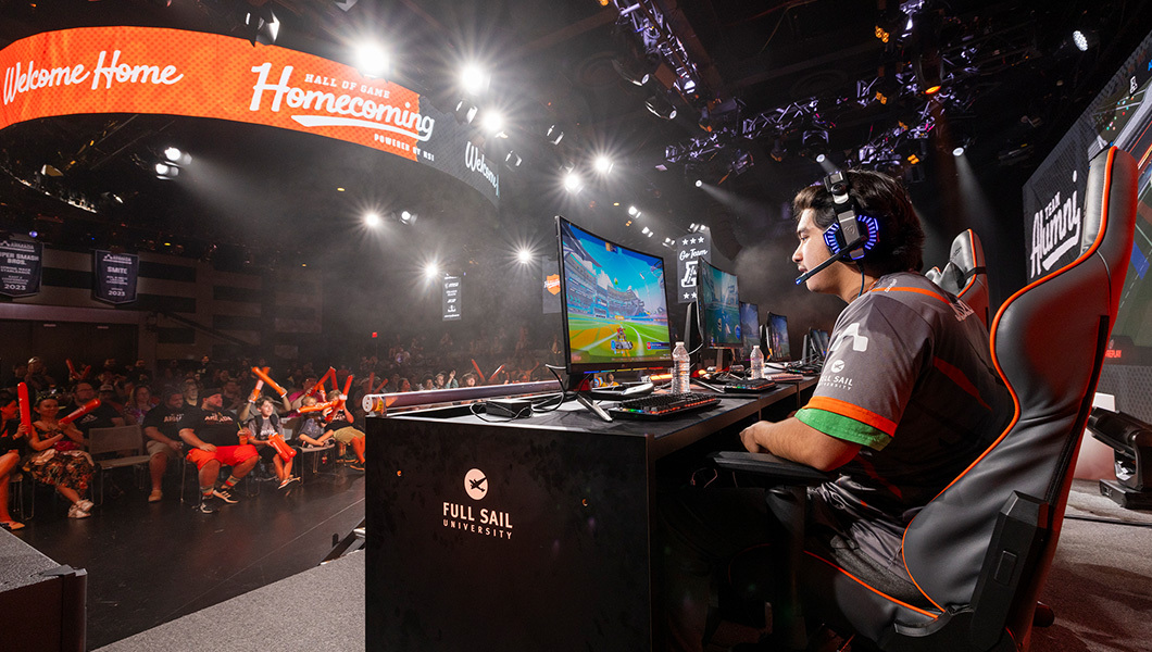 On stage, a student wearing a headset plays a video game on a PC. A large crowd cheers him on from below.