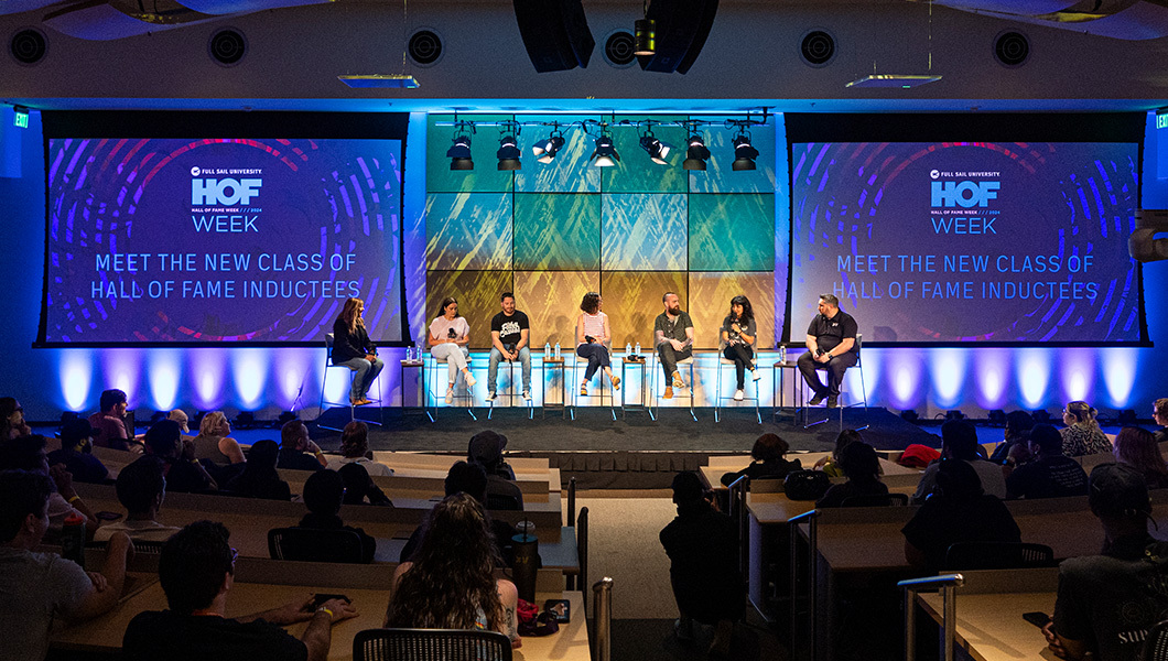 Six people sit on chairs on a stage in front of a sitting crowd. The screen behind them reads “Meet The New Class of Hall of Fame Inductees.”