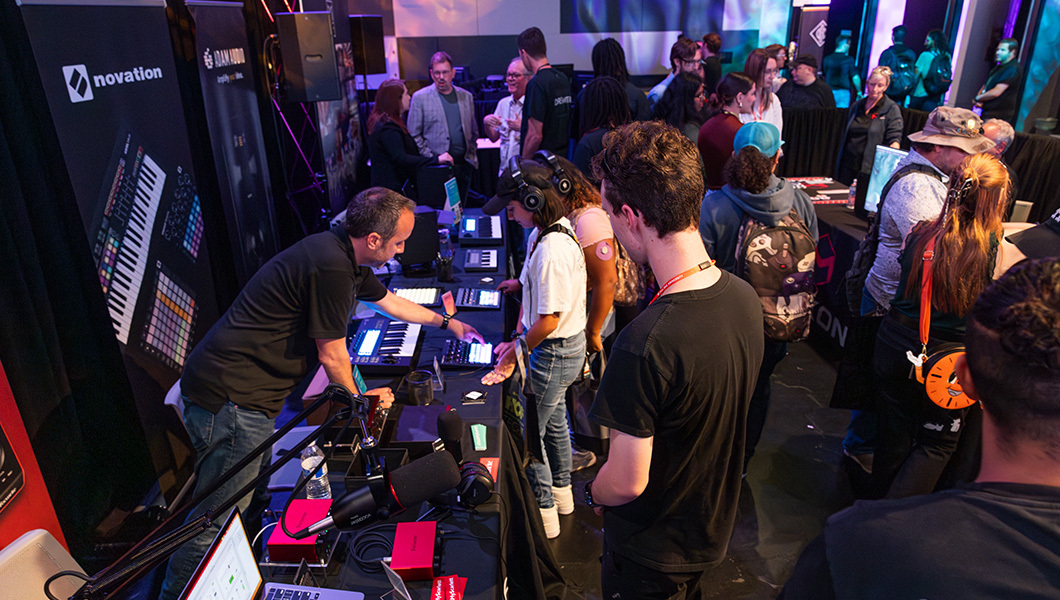 A booth is set up for the company “novation” with many electronics on the table. Behind the booth, a man shows the technology to waiting students. More booths and students mill about in the background.