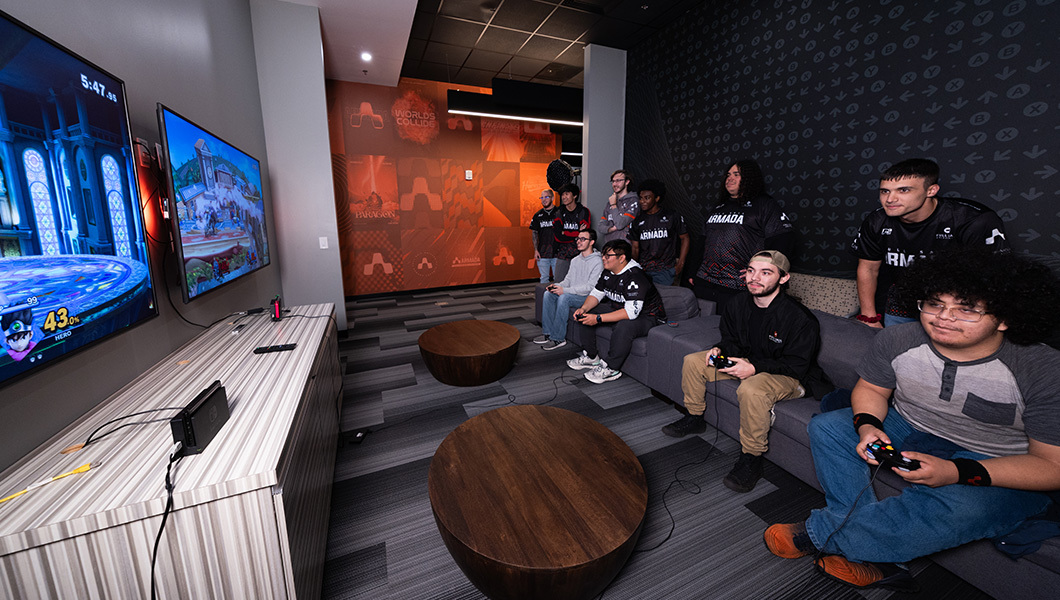 Gamers compete in a lounge with dual screens, a PlayStation 5, and Armada-branded decor, while a crowd watches in the background.