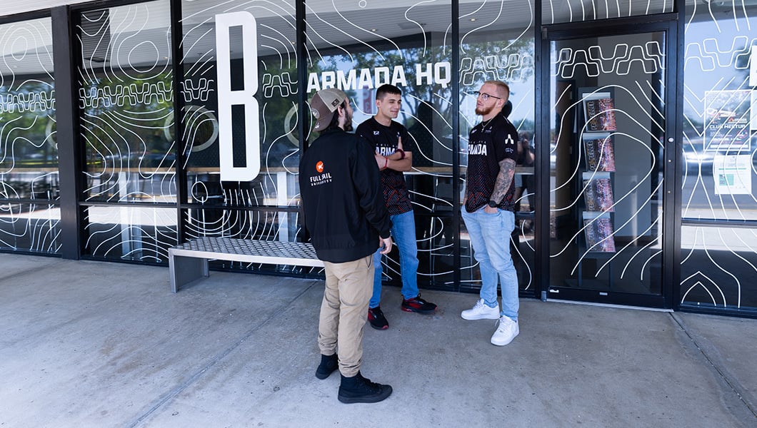 A group of three students is seen standing outside in front of windows with a decal that reads “Armada HQ.”