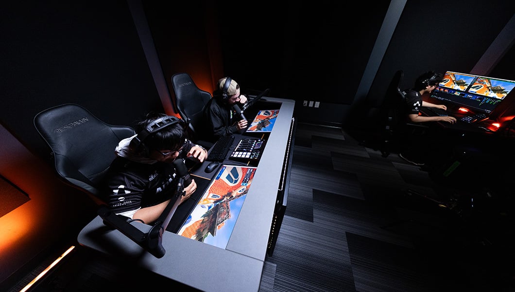 Two students are seated at a broadcast desk, while another is seen in the corner, operating controls on a PC in a dimly lit room.