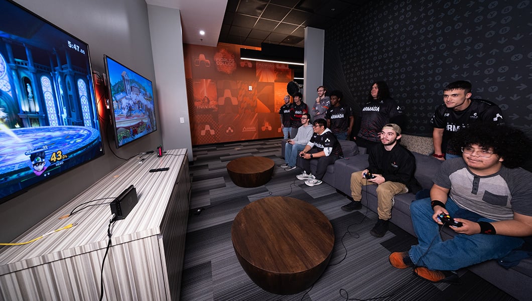 A group of Full Sail students, some in plain clothes and some in Armada attire, are seated on couches and playing console games together.