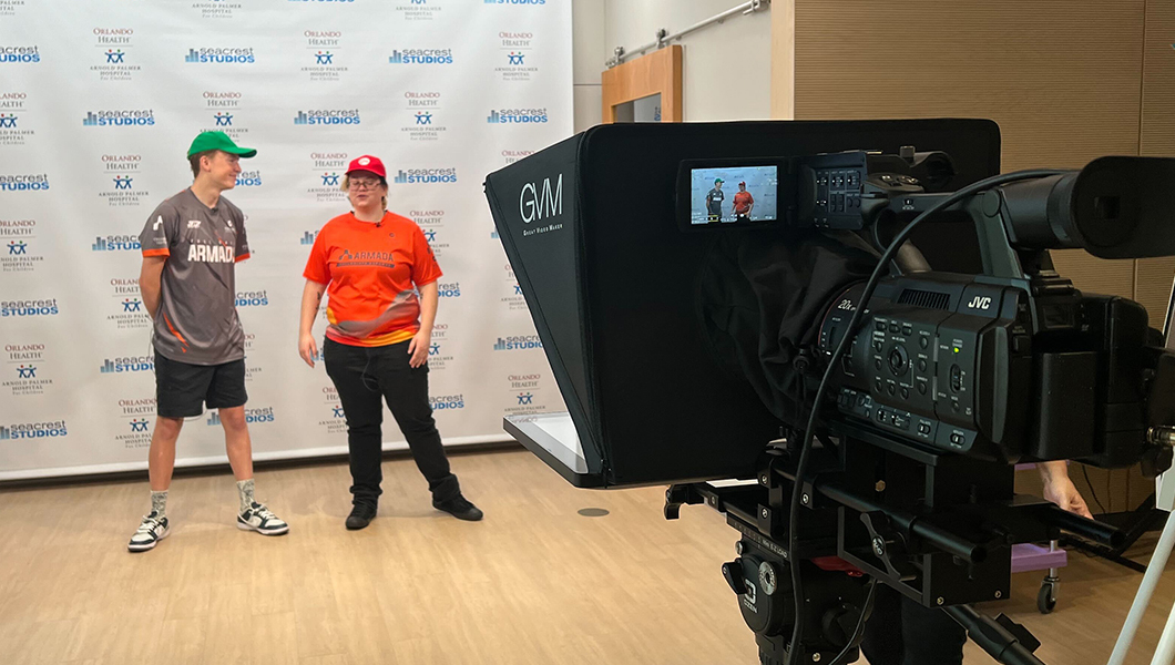 Two people stand in front of a “Seacrest Studios” backdrop, one wearing a green hat and the other a red hat, with a camera filming them.
