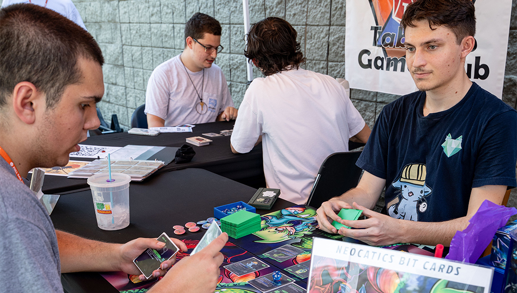 Two students sit across from each other at a table and play a card game. Behind them, another pair of students plays a card game at another table.