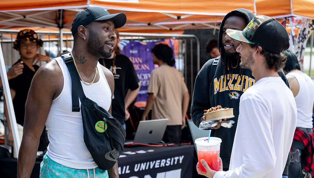 Three students stand and talk to each other in front of orange tents. Two of them wear baseball caps and white shirts, the third wears a hoodie, and one holds a red drink.