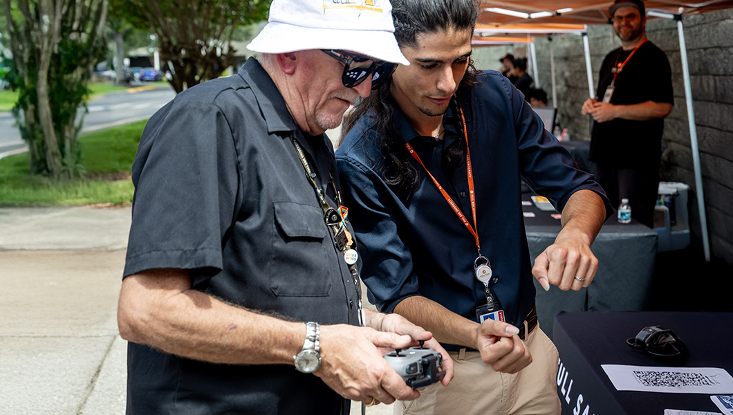 A staff member wears a black shirt and a bucket hat and holds a controller while a student stands next to them and gestures how to use the controller.