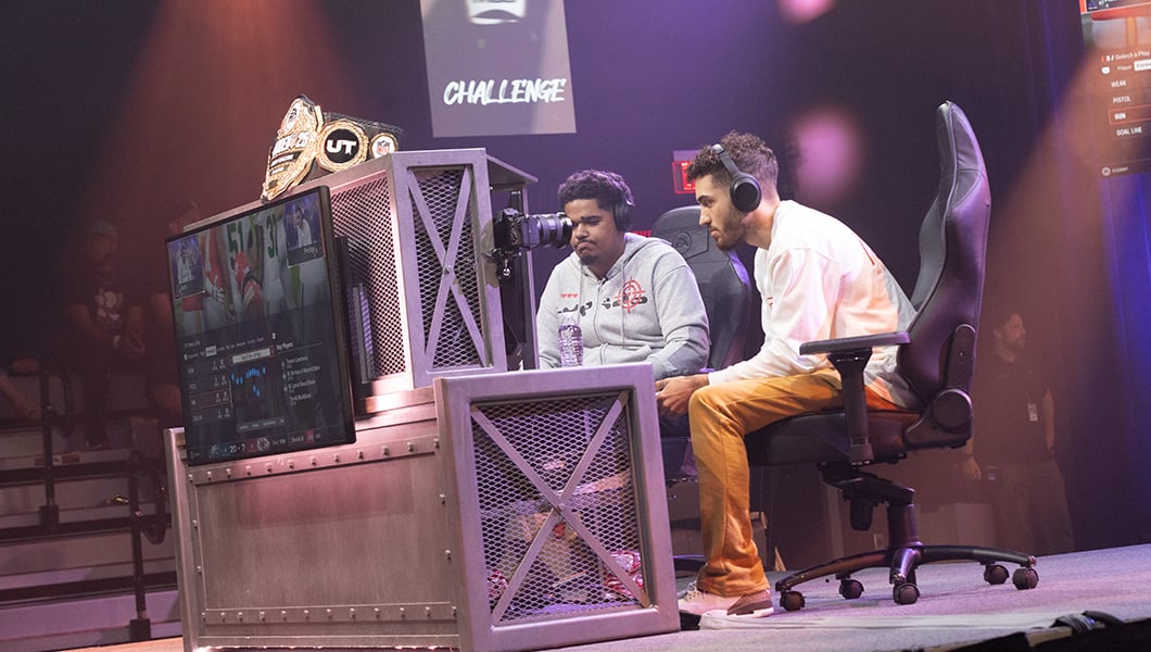 Two focused ‘Madden’ players compete in the Fortress, playing on stage in front of a screen, with a championship belt displayed on the stand.