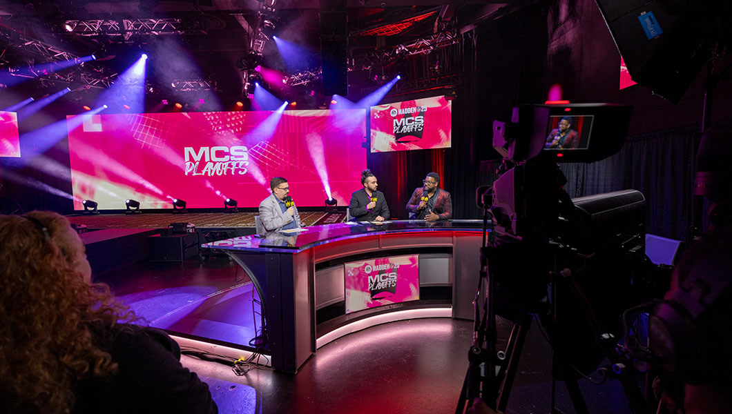 Hosts commentate from a desk in an esports arena featuring "MCS Playoffs" branding, while a camera operator works in the foreground.