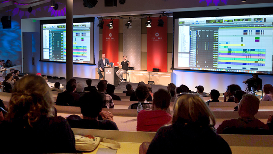 Students attend a live presentation in Full Sail’s Entertainment Business Auditorium featuring large screens and stage lighting.