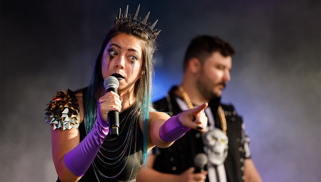 A woman in a metal crown and stage makeup pointing outward while making an exaggerated face at the crowd.