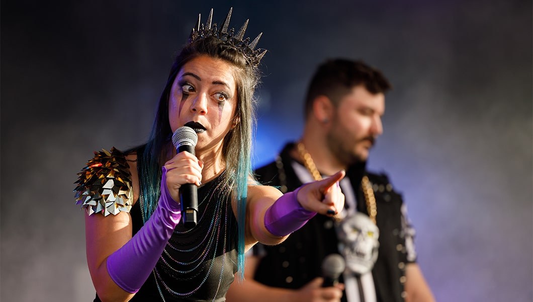 A woman in a metal crown and stage makeup pointing outward while making an exaggerated face at the crowd.