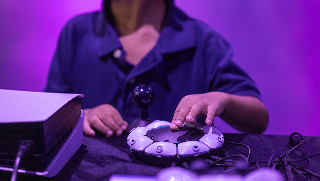 A student sits at a table and presses buttons on a PlayStation 5 Access Controller. Nearby is a PlayStation 5 console and a wireless controller.