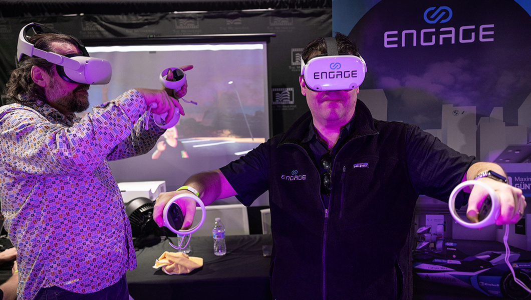 Two men wear virtual reality headsets. They both have their arms extended upward and seem to be enjoying themselves.