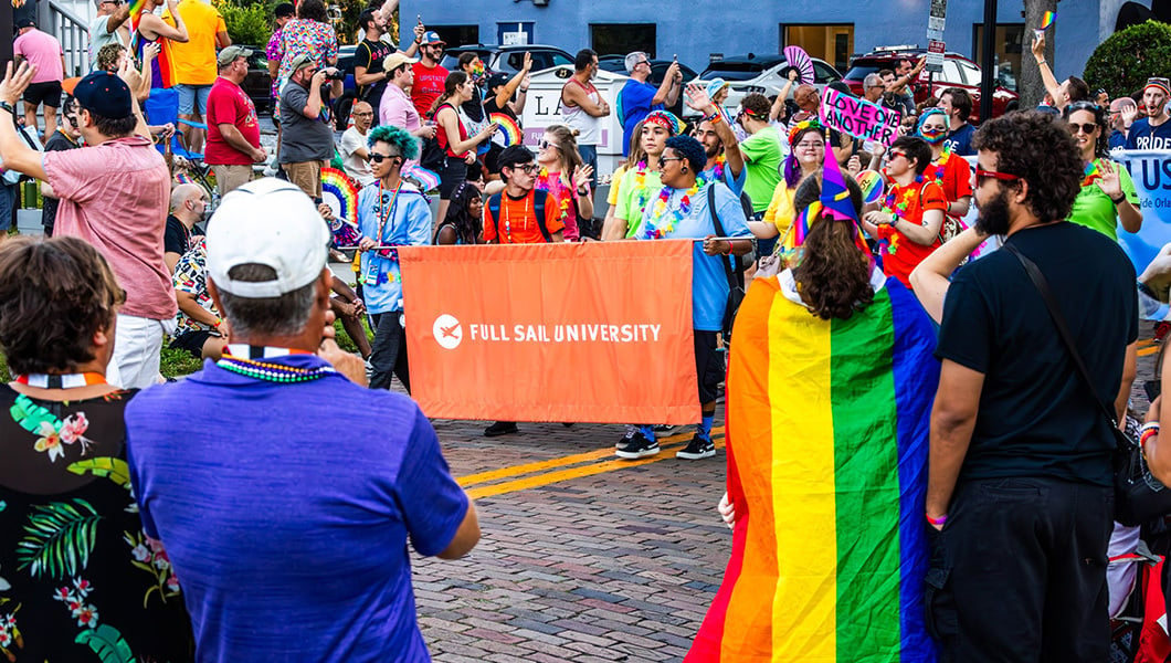 A group of people walking down the street through a parade carrying a large orange banner that reads “Full Sail University.”